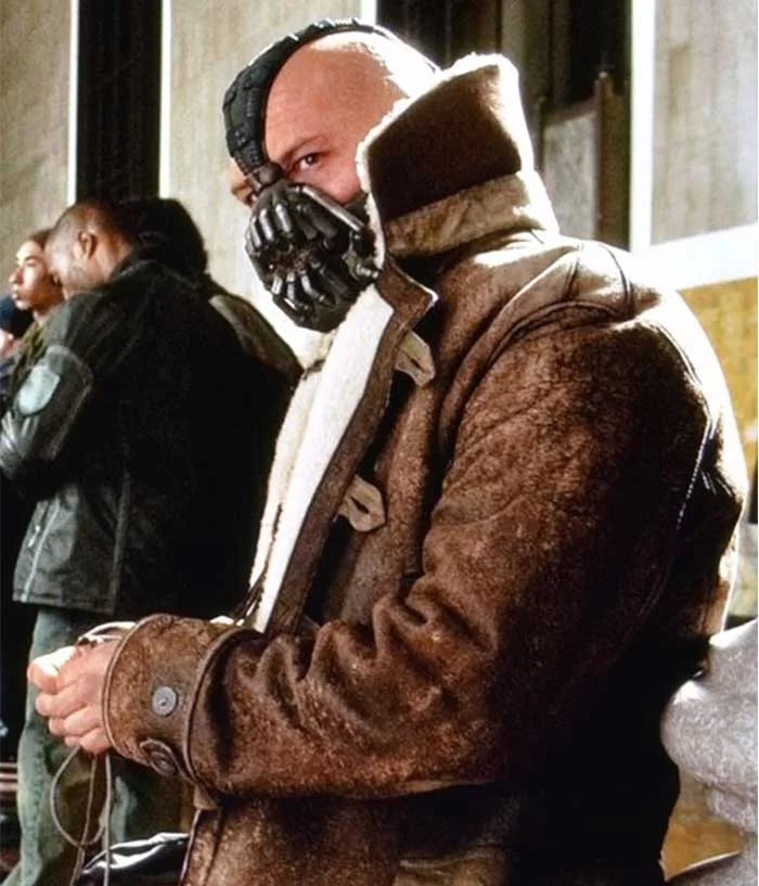A person wearing the Tom Hardy Bane Jacket by Zing Apparel, featuring a brown finish and shearling lining, along with a black mask, sits indoors in a crowded space, looking to the side.