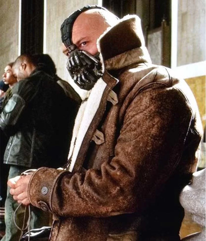 A person wearing the Tom Hardy Bane Jacket by Zing Apparel, featuring a brown finish and shearling lining, along with a black mask, sits indoors in a crowded space, looking to the side.