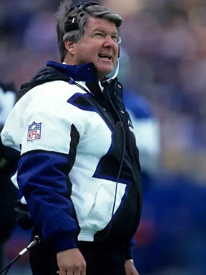 A football coach stands on the sideline during a game, wearing the Jimmy Johnson Cowboys Jacket by Zing Apparel—a multicolor cotton-polyester NFL jacket with a hooded collar and a headset on, looking up.