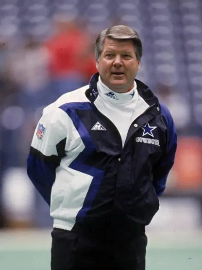 A football coach stands on the field in a Jimmy Johnson Cowboys Jacket by Zing Apparel, featuring a multicolor blue and white design with a hooded collar. Blurred stadium seats are visible in the background.