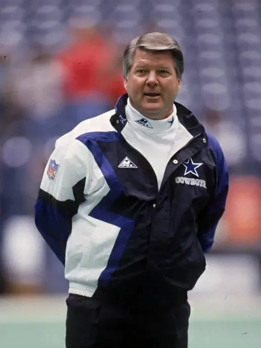 A football coach stands on the field in a Jimmy Johnson Cowboys Jacket by Zing Apparel, featuring a multicolor blue and white design with a hooded collar. Blurred stadium seats are visible in the background.