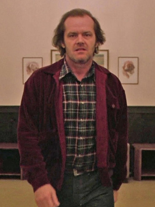A man with medium-length hair wears the Zing Apparel "The Shining Jack Torrance Corduroy Jacket" featuring a shirt-style collar, over a plaid shirt and jeans. Indoors, he looks serious; three framed pictures hang on the wall behind him.