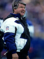 A football coach stands on the sideline during a game, wearing the Jimmy Johnson Cowboys Jacket by Zing Apparelâ€”a multicolor cotton-polyester NFL jacket with a hooded collar and a headset on, looking up.