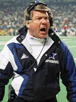 A football coach in a Jimmy Johnson Cowboys Jacket by Zing Apparel with a hooded collar and a headset stands on the sidelines, shouting during a game, with a blurred stadium crowd in the background.