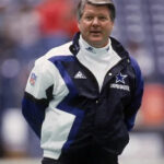 A football coach stands on the field in a Jimmy Johnson Cowboys Jacket by Zing Apparel, featuring a multicolor blue and white design with a hooded collar. Blurred stadium seats are visible in the background.