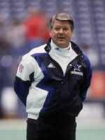 A football coach stands on the field in a Jimmy Johnson Cowboys Jacket by Zing Apparel, featuring a multicolor blue and white design with a hooded collar. Blurred stadium seats are visible in the background.